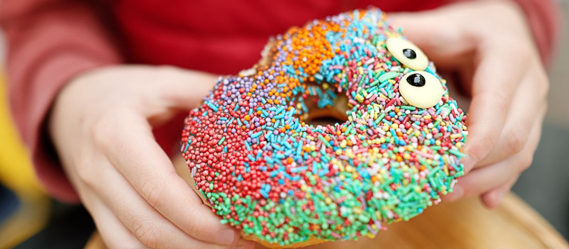 Little child eating funny colorful donut in a street cafe while walking in the city center. Sweet pastries and fastfood for children. Unhealthy nutrition for kids.