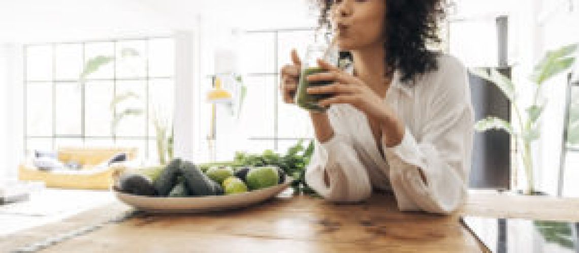Young african american woman drinking green juice with reusable bamboo straw in loft apartment. Home concept. Healthy lifestyle concept. Copy space